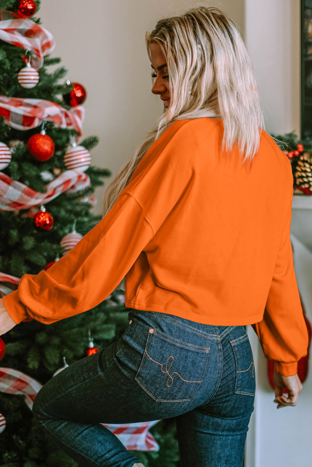 A woman with blonde hair, wearing an Orange Game Day Lettering Rugby Football Notched Neck Sweatshirt and blue jeans, stands next to a decorated Christmas tree.