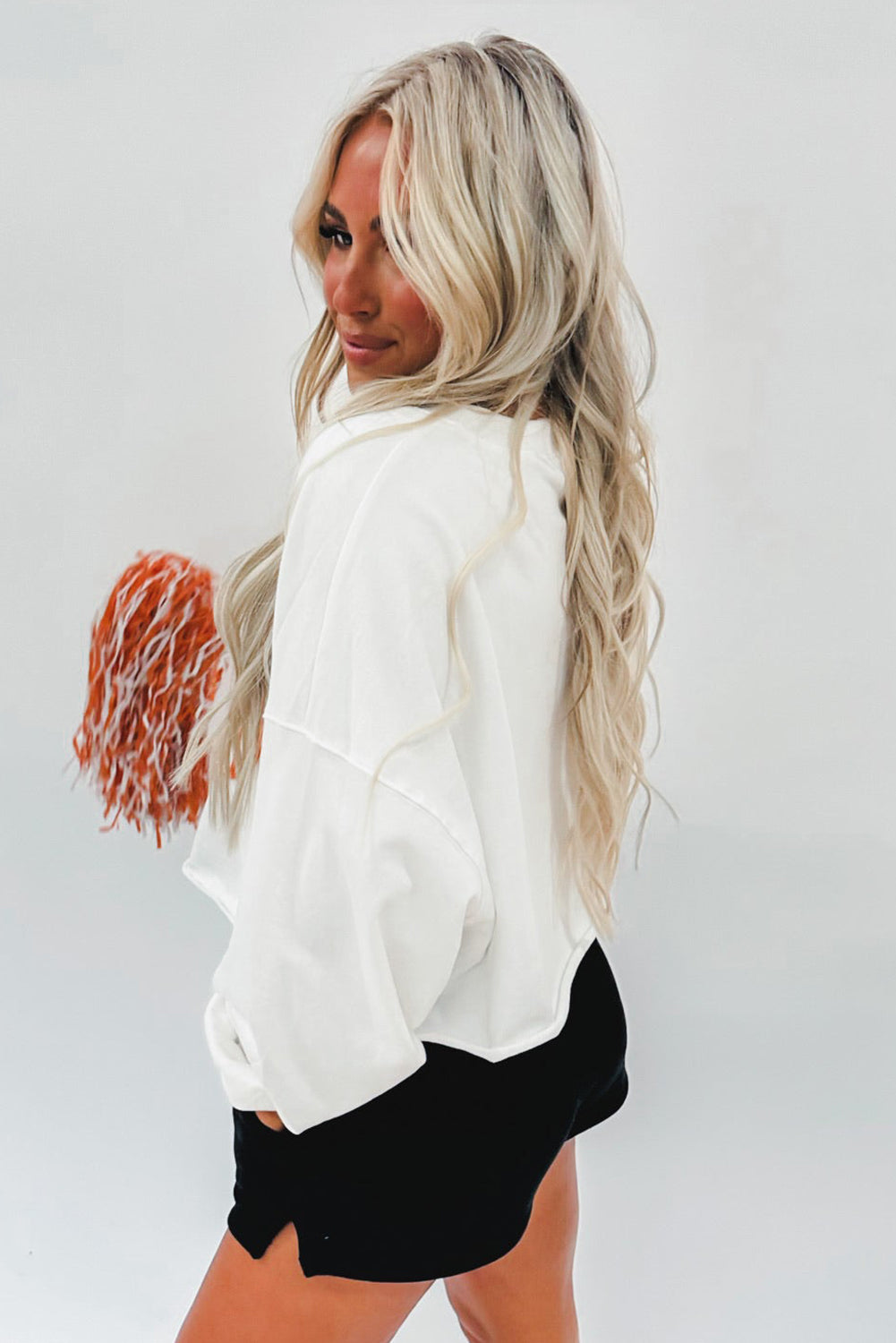 A woman with long blonde hair dressed in a chic White Game Day Lettering Rugby Football Notched Neck Sweatshirt and blue jeans stands near a Christmas tree adorned with red and white ornaments and ribbon.