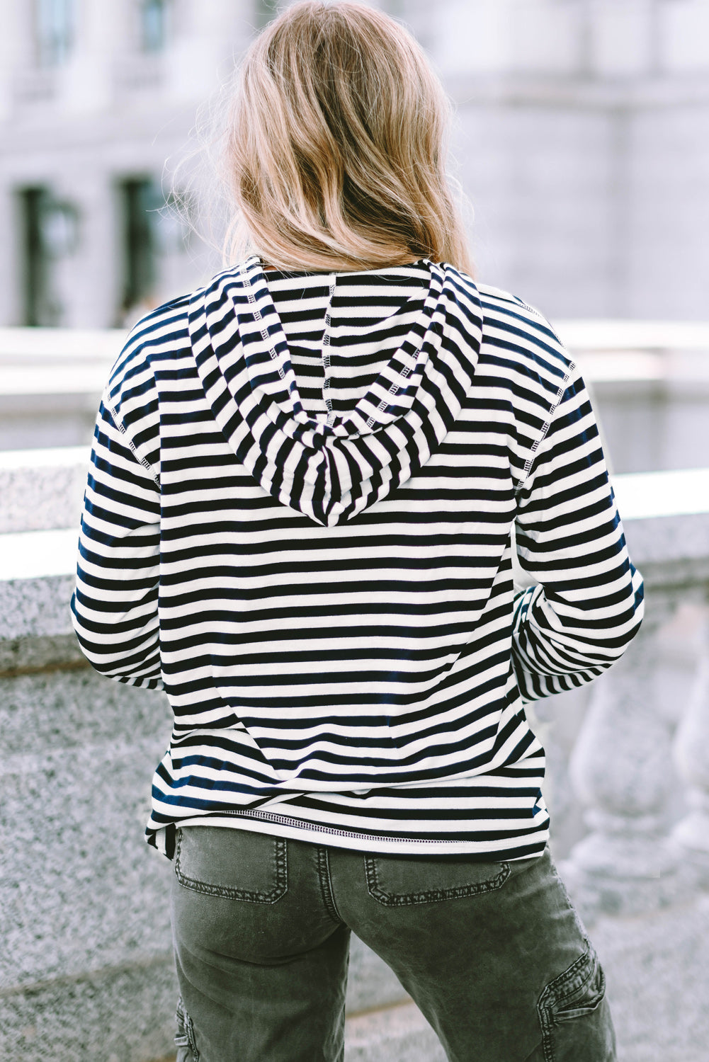 A person with long, blonde hair wearing a trendy Stripe Kangaroo Pocket Drawstring Hoodie with black and white stripes and dark green pants is standing outdoors, facing away from the camera.