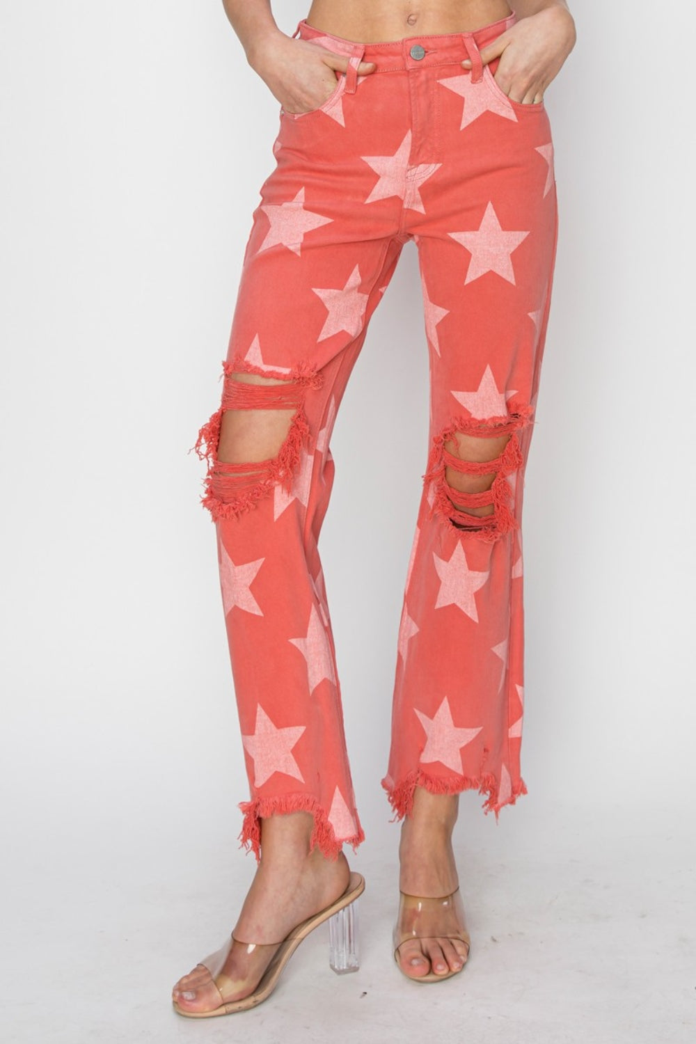 A person wearing the RISEN Full Size Distressed Raw Hem Star Pattern Jeans in a coral color, complete with distressed rips at the knees and raw frayed hems. The rock 'n' roll style is completed with clear high-heeled sandals.