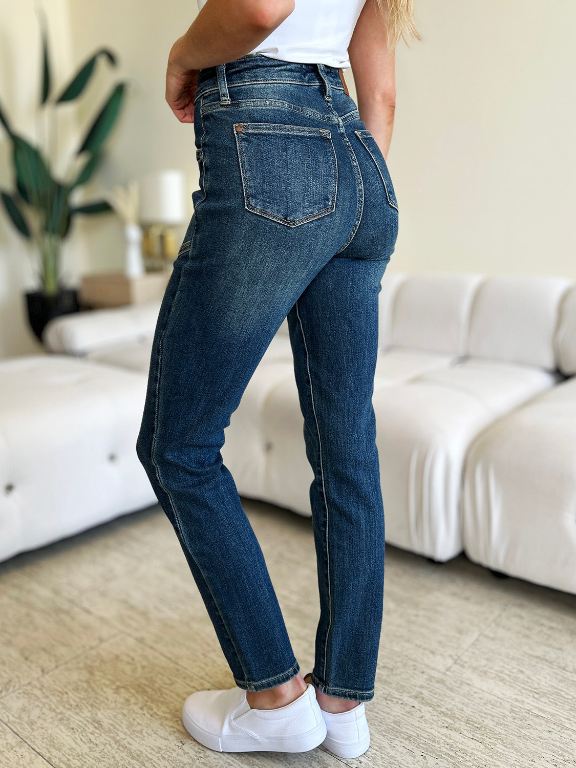 A person wearing Judy Blue Full Size High Waist Skinny Jeans made of stretchy denim fabric stands in a room with light-colored furniture and wooden flooring, paired perfectly with white slip-on shoes.