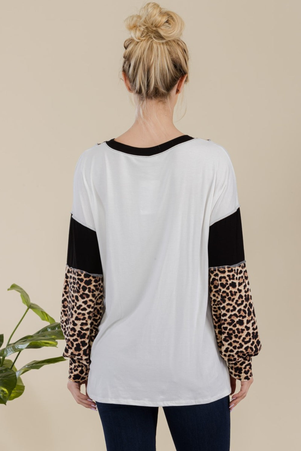 A woman stands in front of a simple background with a plant, wearing the Celeste Full Size Leopard Color Block Exposed Seam T-Shirt, which boasts leopard print cuffs and striking black and white color blocks.