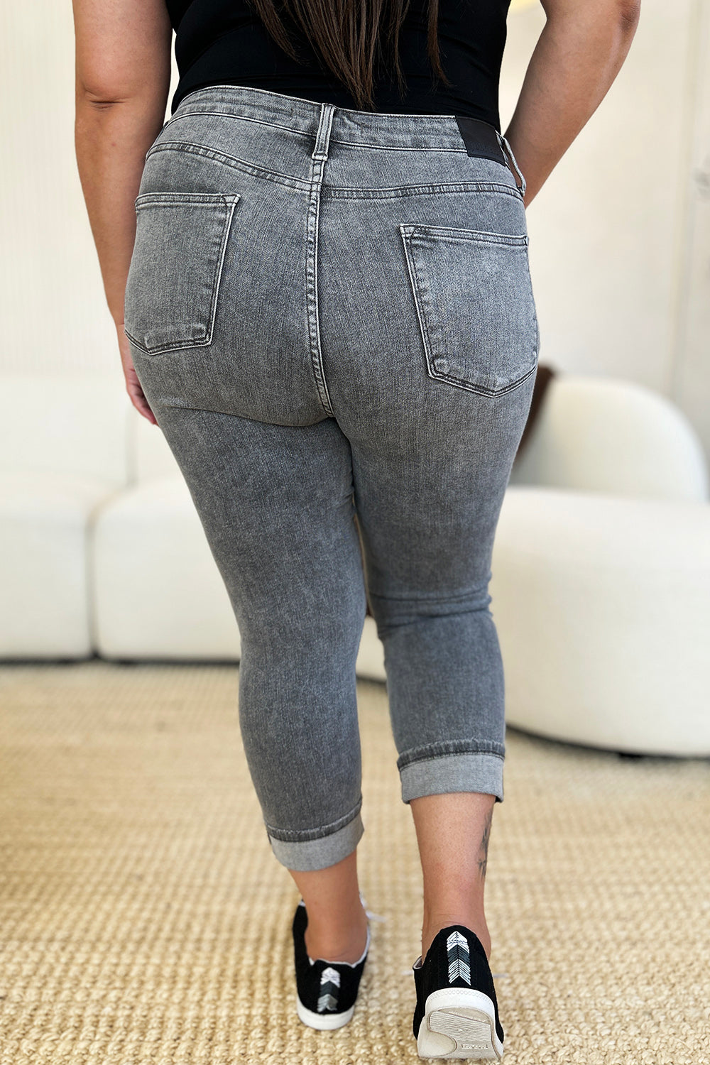 A person wearing Judy Blue Full Size Button Fly High Waist Cuffed Capris in gray, paired with a white top and white sneakers, stands on a beige carpeted floor.