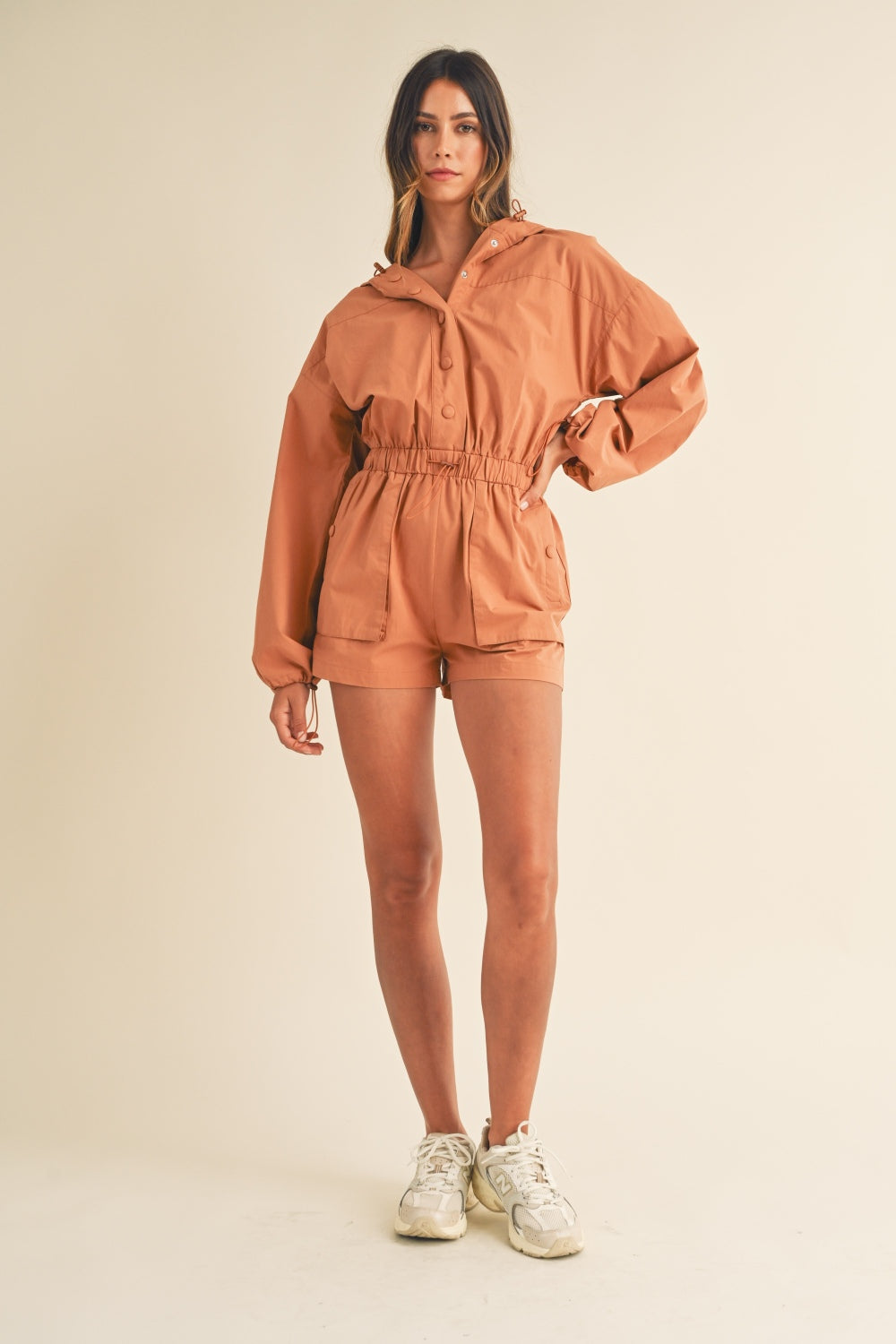 A person wearing the Mable Snap Down Long Sleeve Hooded Romper, featuring a brown color, long sleeves, a snap-down front, and an elastic waistband, is standing against a plain background.