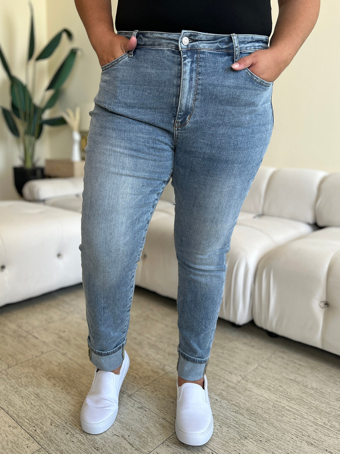 A person wearing Judy Blue Full Size High Waist Cuff Hem Jeans and white slip-on shoes stands in a living room with a white couch and plants in the background.