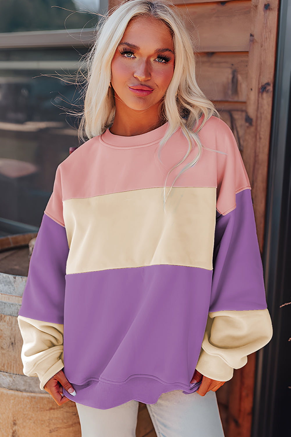 A woman with long blonde hair stands with her back to the camera, wearing a trendy Meadow Mauve Colorblock Patchwork Drop Shoulder Sweatshirt. She is next to a wooden barrel and large pampas grass decorations.