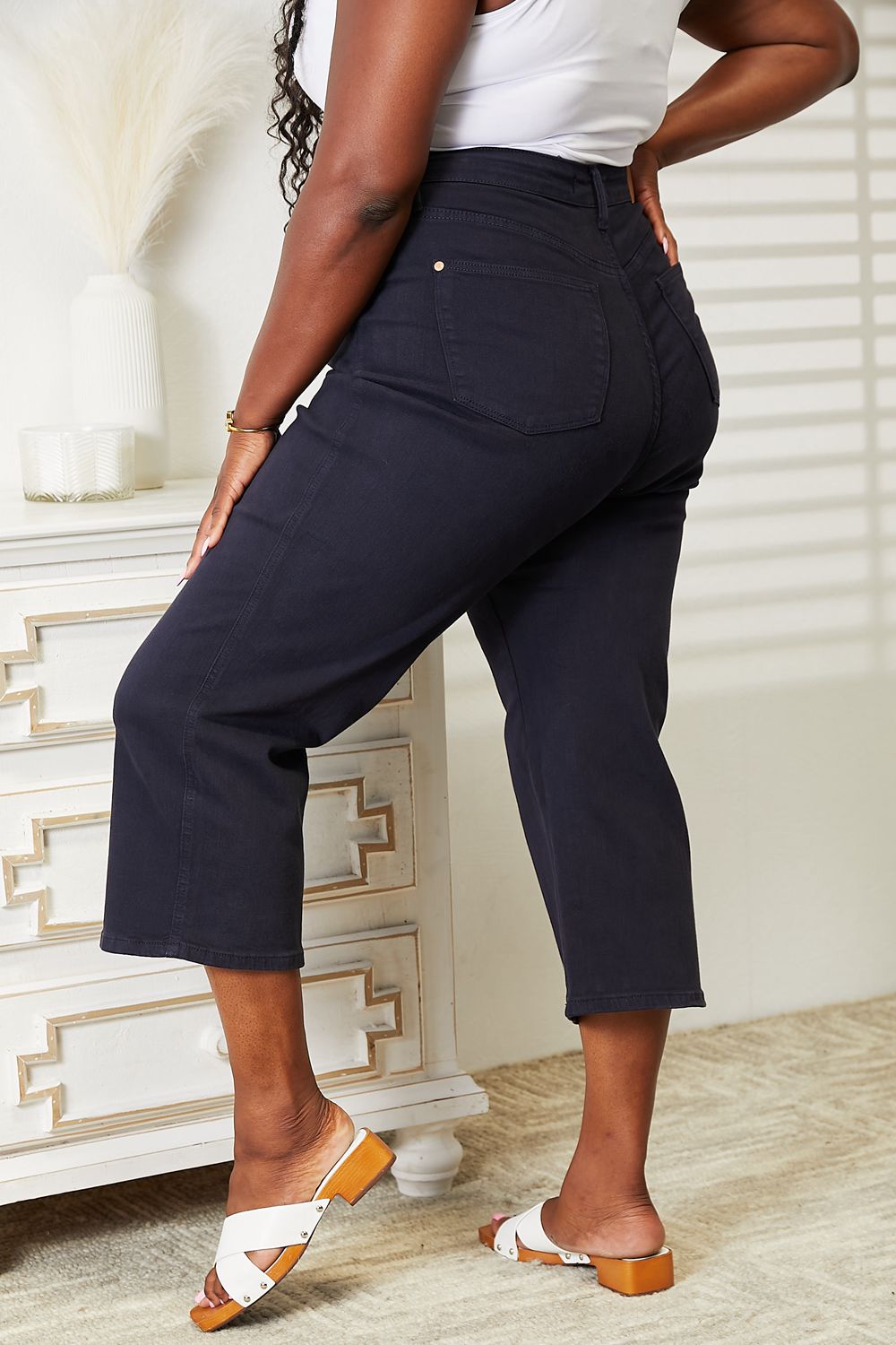 A person wearing a white sleeveless top and Judy Blue Full Size High Waist Tummy Control garment-dyed wide cropped jeans in black stands indoors next to a white dresser.