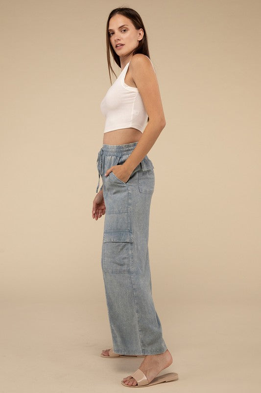 A woman is wearing Washed Linen Elastic Band Waist Cargo Pants in light blue, featuring wide legs, front pockets, an elastic waistband, and a drawstring closure. She is standing with her hands in her pockets, paired with a white top and beige slide sandals.