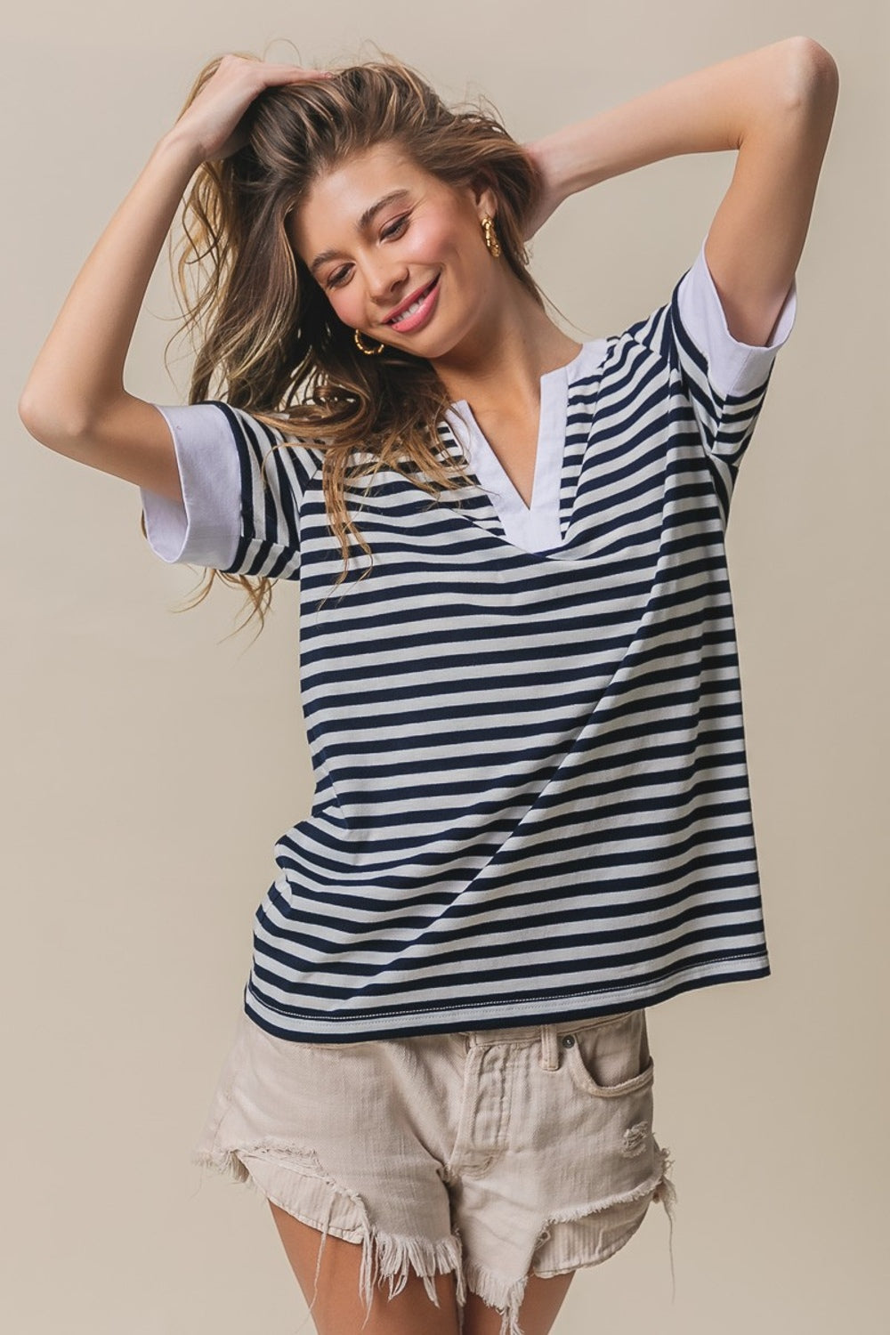 A person with long hair, wearing a BiBi Contrast Striped Notched Knit Top in navy and white and beige shorts, is posing with one hand in their hair, smiling against a beige background.