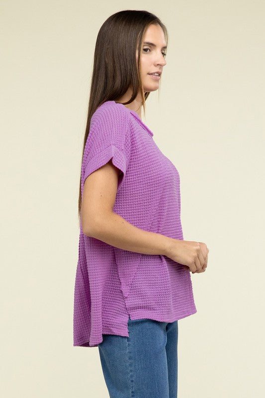 A woman with long brown hair is wearing a purple Brushed Waffle Exposed-Seam Short Sleeve Top and blue jeans, standing against a plain beige background. This casual wardrobe choice, with its short sleeves and side slits, is perfect for everyday wear.