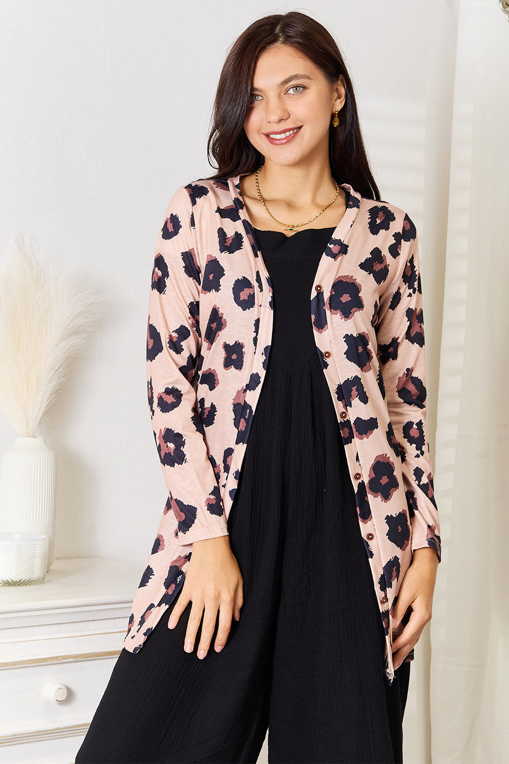 A woman wearing the Double Take Printed Button Front Longline Cardigan, featuring a pink floral pattern, over a black dress, stands indoors near a white vase with dried plants.