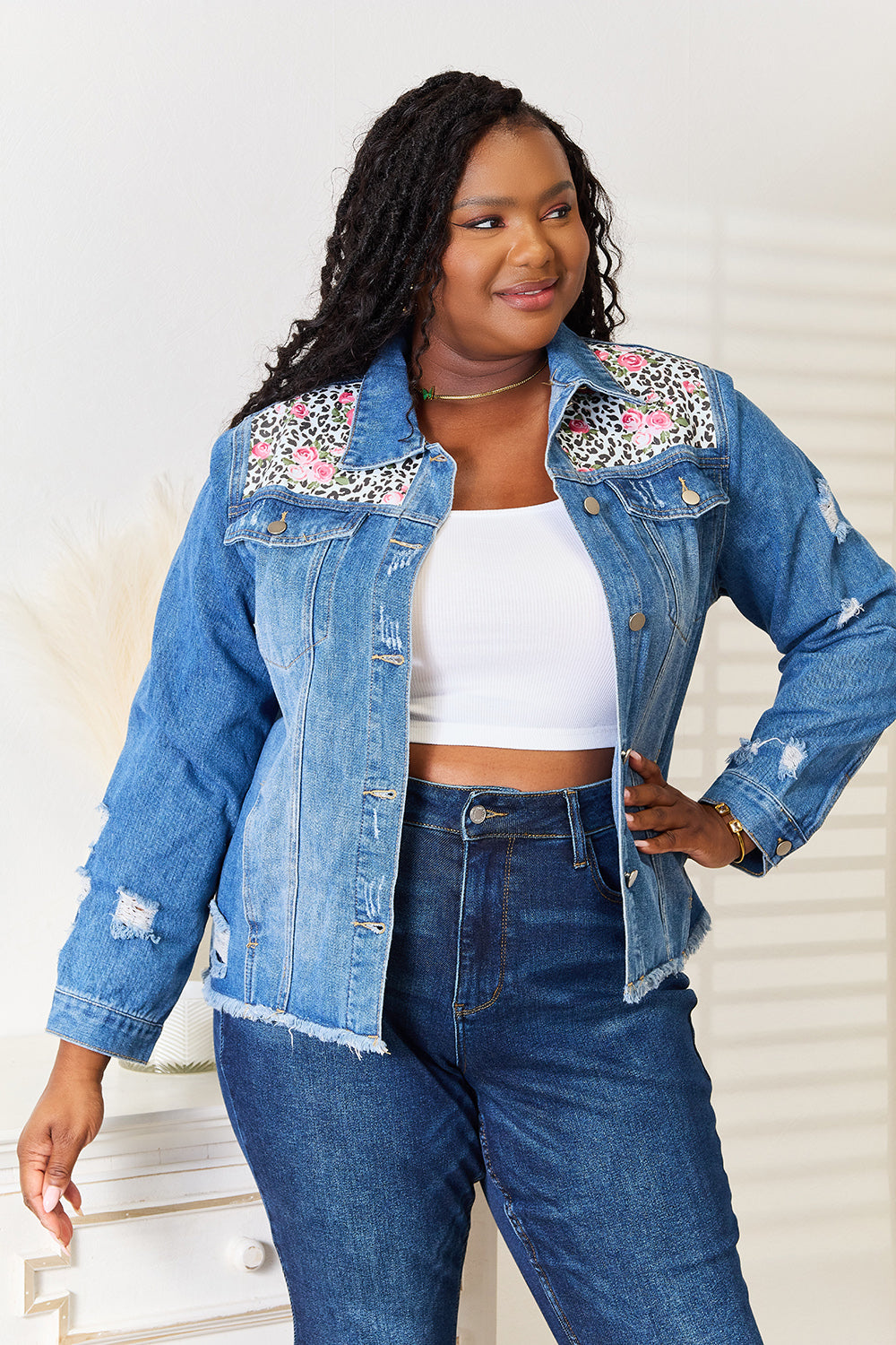 A person smiles while wearing the Baeful Leopard Floral Distressed Raw Hem Denim Jacket with embroidered patches, paired with a white shirt and blue jeans, as they stand indoors near a white piece of furniture.