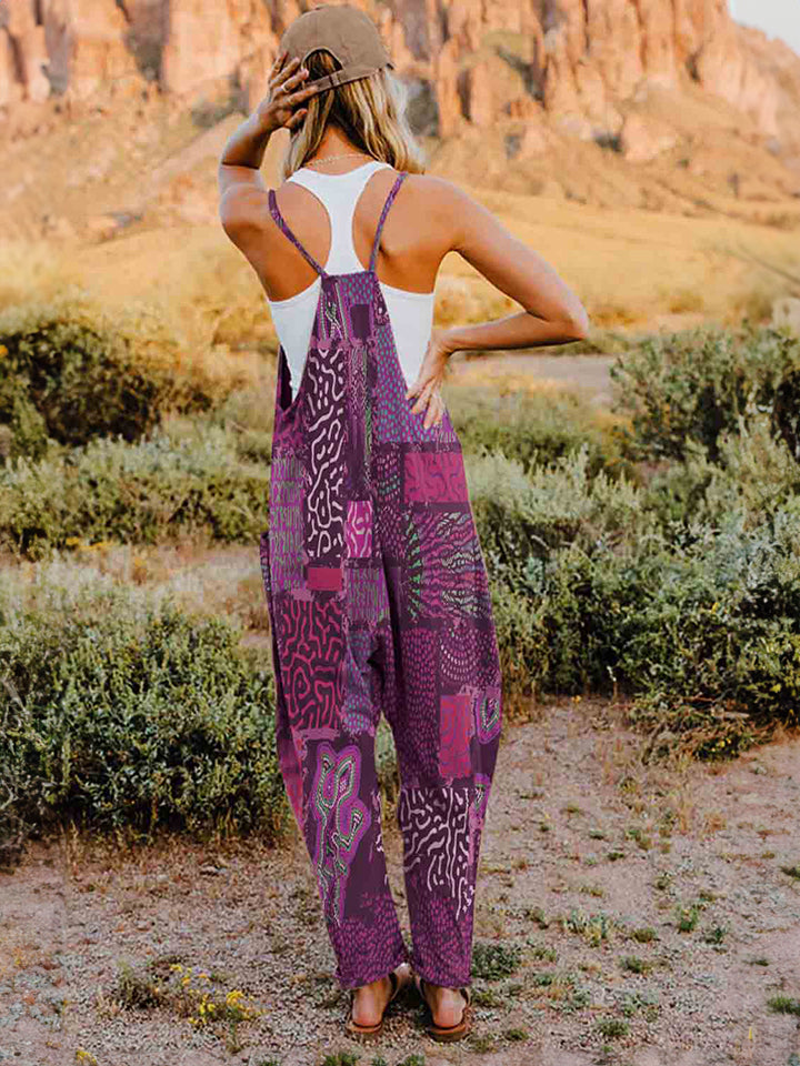 A person standing outdoors in front of rocky terrain, wearing a Full Size Printed V-Neck Sleeveless Jumpsuit and a hat.