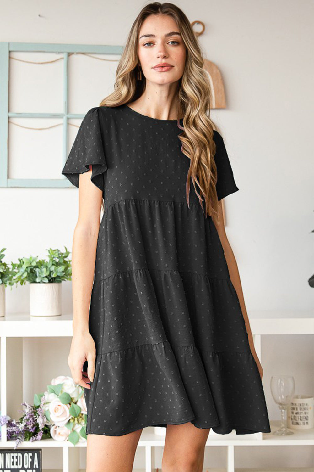 A woman elegantly poses indoors wearing the Heimish Swiss Dot Short Sleeve Tiered Dress in chic black, showcasing its stylish tiered design. The lush plants and tidy shelves create a perfect backdrop for her ensemble.