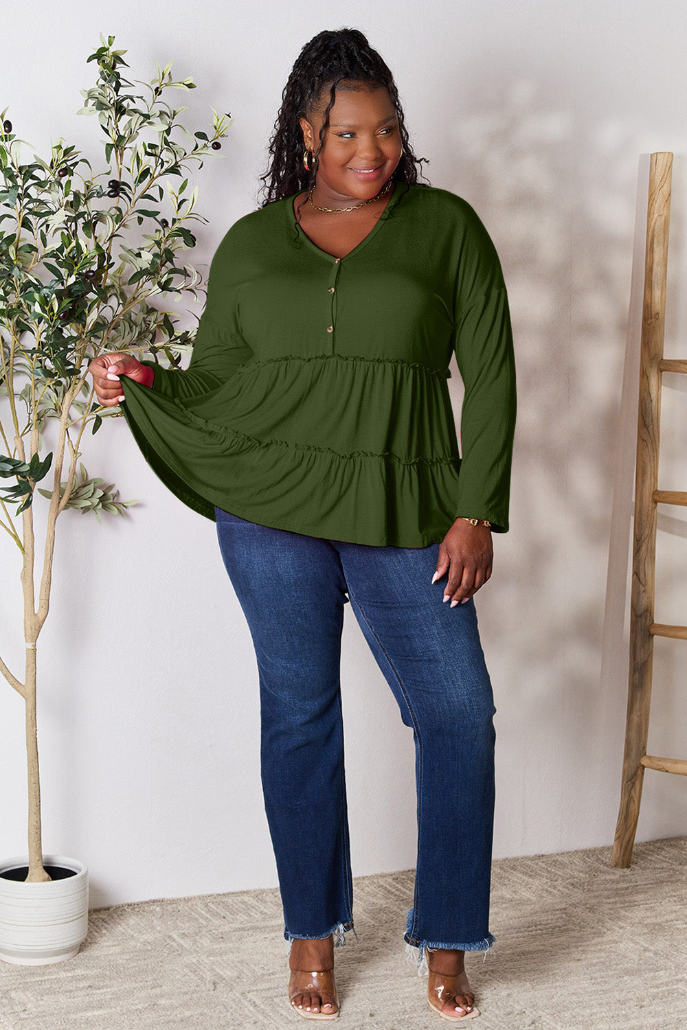 Person stands indoors near a plant, wearing the Double Take Half Button Long Sleeve Ruffle Hem Blouse in gray and blue jeans, smiling while looking to the side. The slightly stretchy fabric of the blouse adds comfort while its buttoned design offers a touch of elegance.