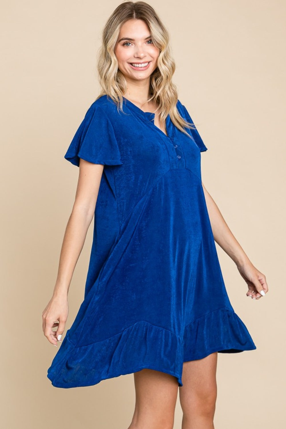 A woman wearing the Culture Code Full Size Short Sleeve Ruffled Asymmetric Hem Dress poses against a beige background.