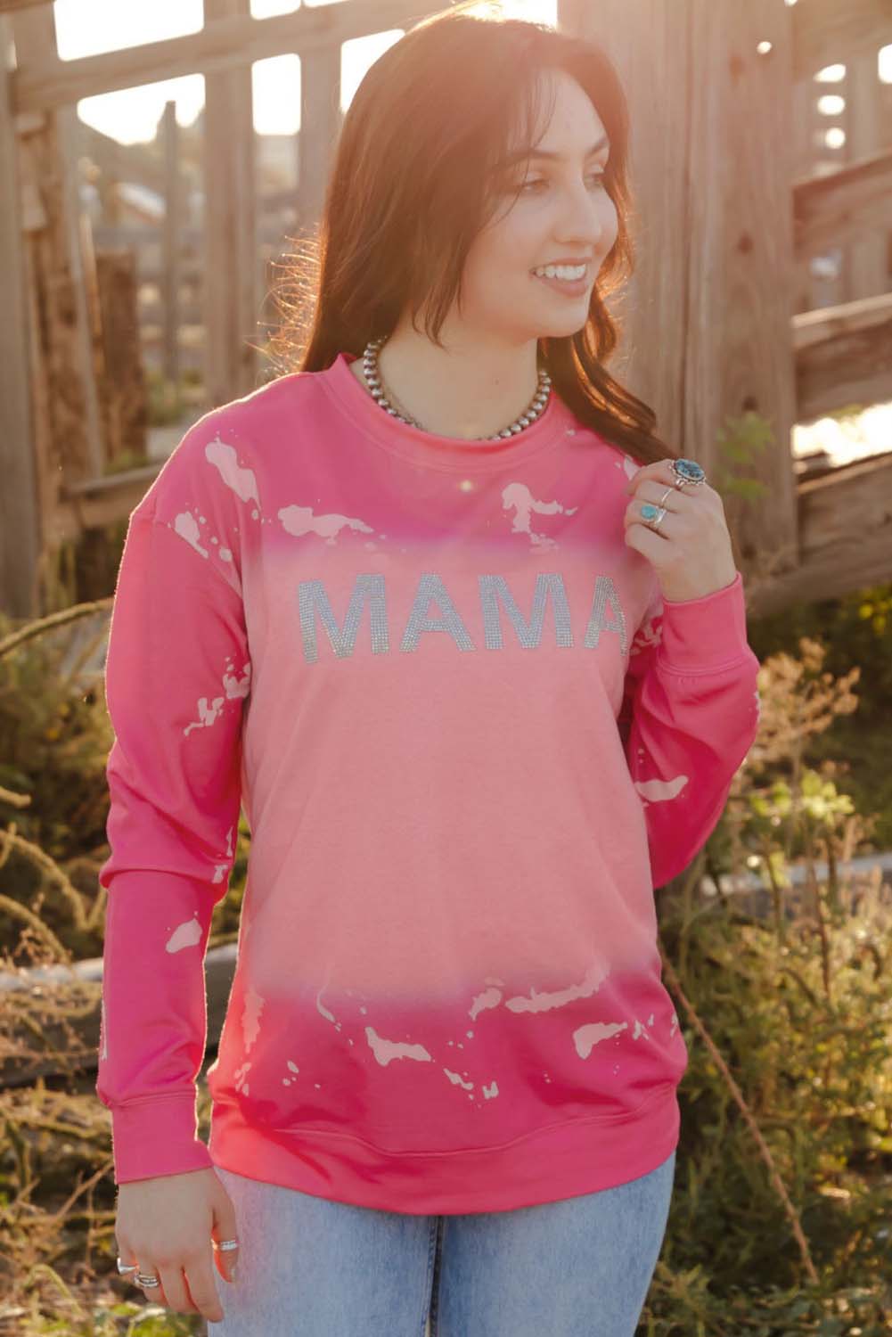 A person with long dark hair is standing outdoors, facing away from the camera, wearing a Pink Mama Rhinestone Letter Graphic Tie Dye Pullover and light blue jeans. Old wooden structures are visible in the background.