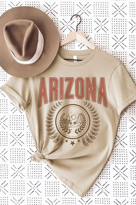 A person wearing a light gray ARIZONA Graphic Tee that showcases a desert scene with cacti and a sun, encircled by a laurel wreath design. This tee is made from 100% cotton and is available in unisex sizing.