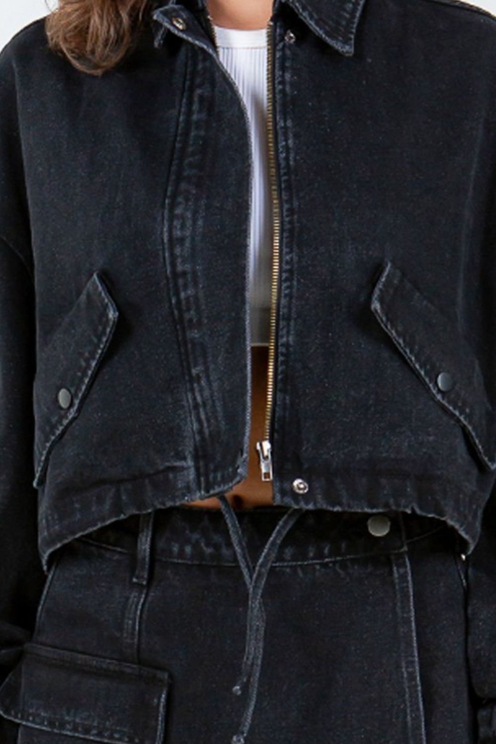 A person with wavy hair is wearing an American Bazi Drawstring Waist Crop Denim Jacket in black, featuring a zippered front closure, paired with a matching black denim skirt.