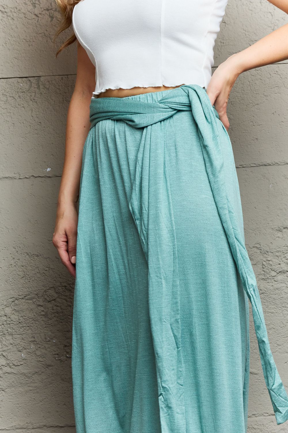 A woman stands against a textured gray wall, wearing the Ninexis Know Your Worth Criss Cross Halter Neck Maxi Dress in teal, featuring a midriff cutout and criss-cross pattern. She pairs it with tan heeled sandals and poses with her right hand on her hip.