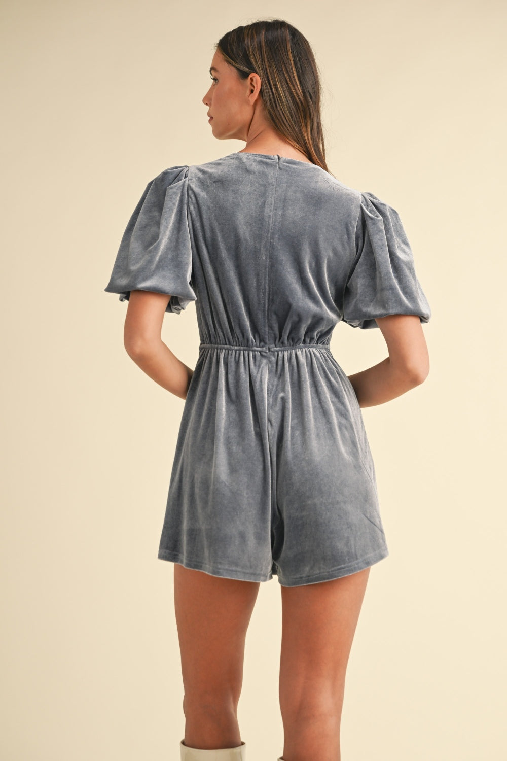 A woman stands against a plain background wearing the Mable Jewel Bow Decor Puff Sleeve Velvet Romper in grey, paired with white boots. The romper, adorned with three jewel bow decor accents on the front, exudes understated elegance.