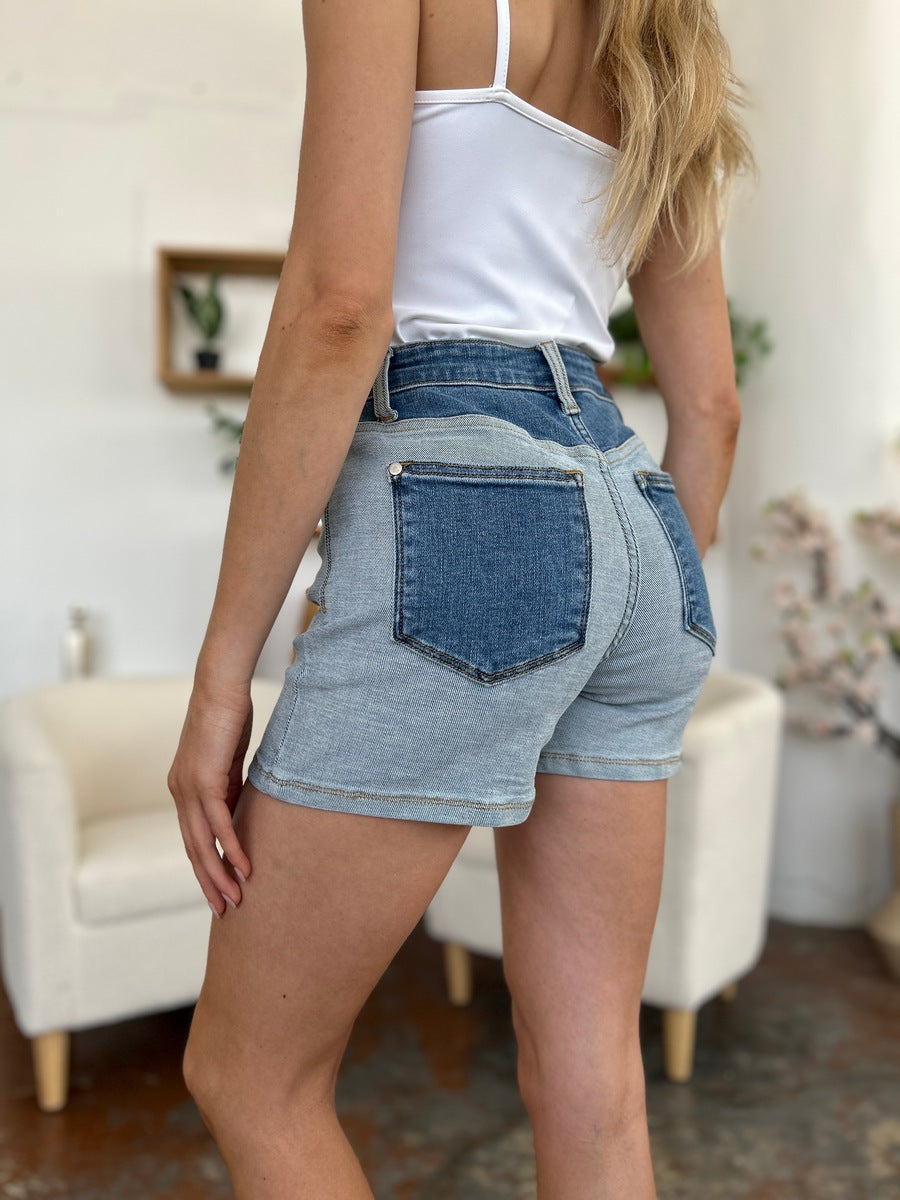 A person models indoors beside a beige chair and a plant, sporting a white t-shirt with the Judy Blue Full Size Color Block Denim Shorts for an effortlessly bold, eye-catching look.