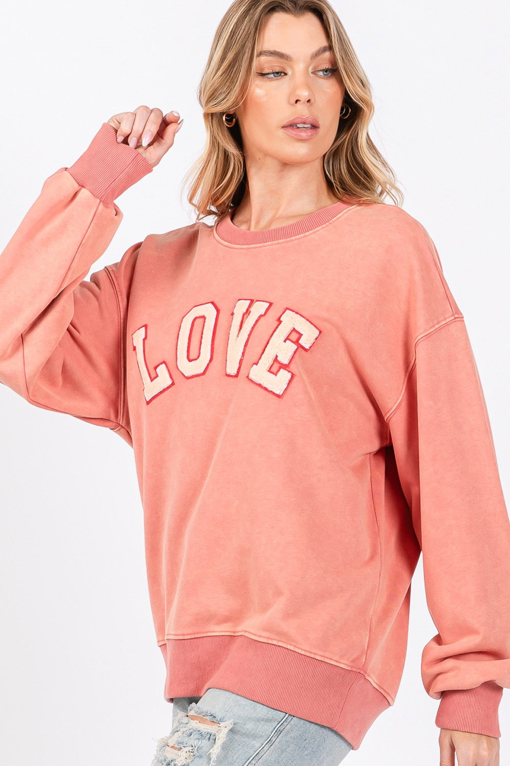 A person with long hair wears a cap, an oversized pink SAGE + FIG LOVE Path Applique Drop Shoulder Sweatshirt, and ripped jeans, standing against a plain background.