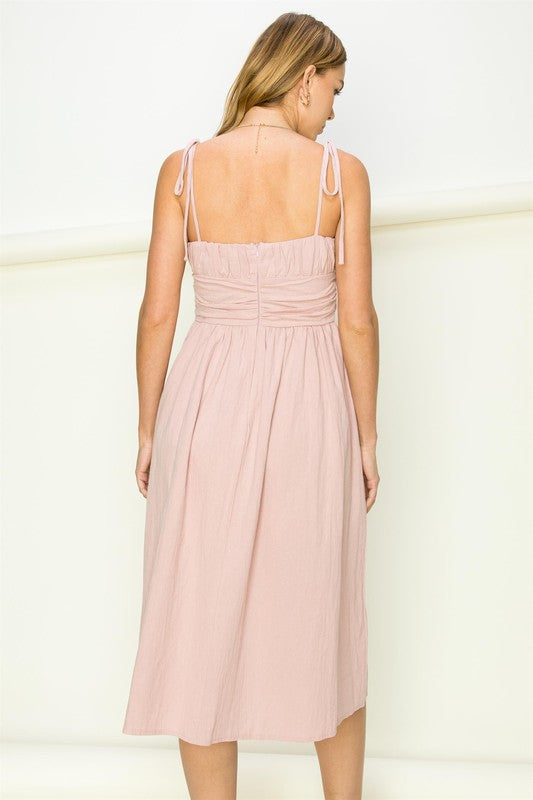 A woman wearing the Get a Clue Tie-Strap Midi Dress, featuring a light pink hue and tie-straps, showcases chic fashion and a flowy silhouette as she stands against a plain background.