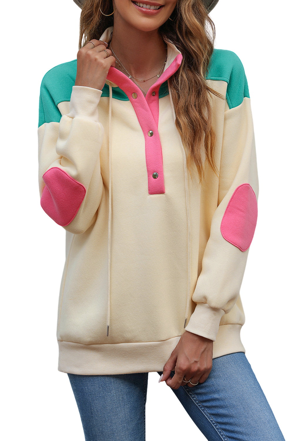 A woman wearing the Apricot Color Block Elbow Patch Half Button Sweatshirt, featuring cream and mint colors with pink elbow patches and a turn-down collar with snap button closure, stands outdoors. She has long wavy hair, sunglasses, a hat, and is wearing blue jeans.