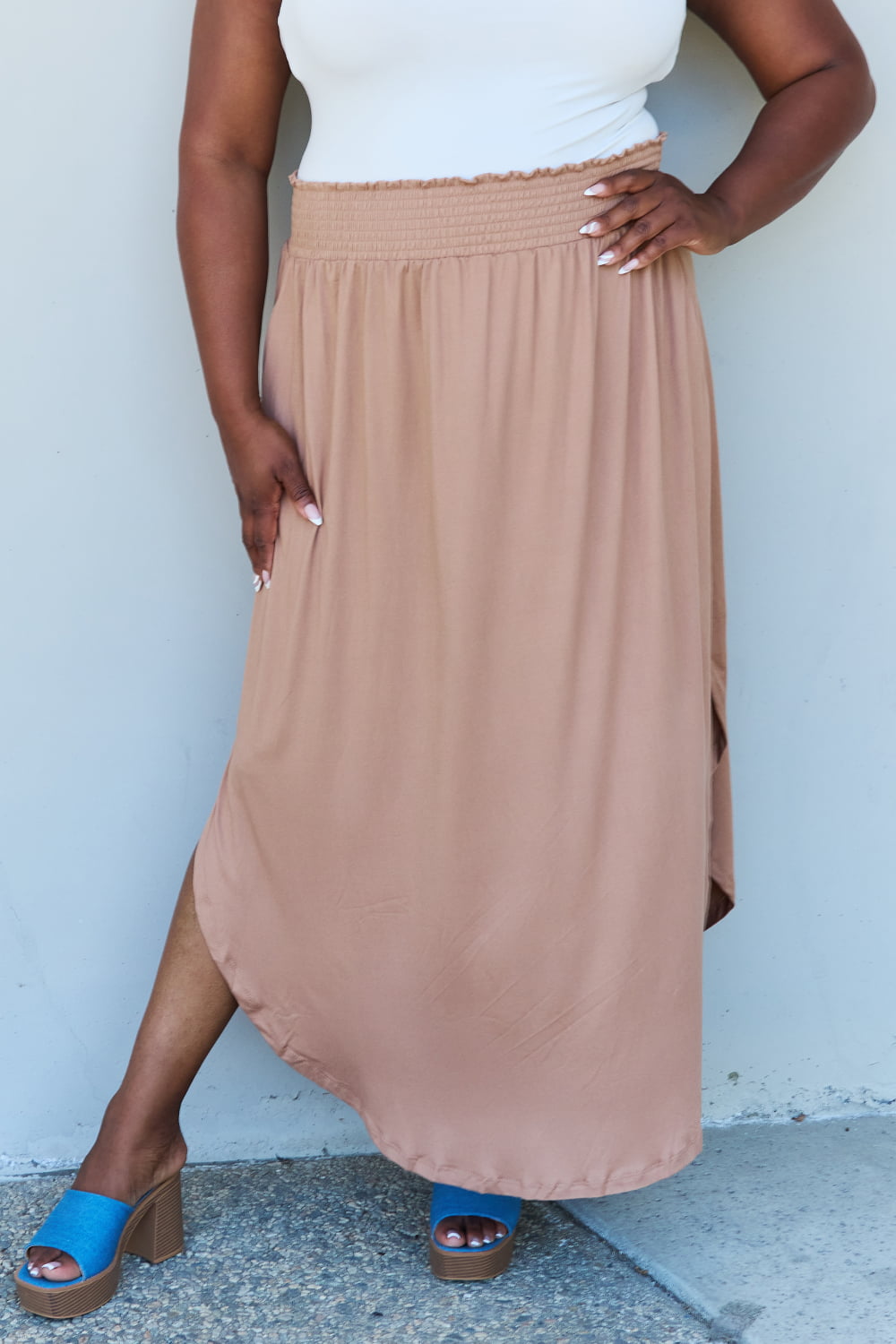 A person stands against a wall, wearing a white sleeveless top, the Doublju Comfort Princess Full Size High Waist Scoop Hem Maxi Skirt in Tan, and blue platform sandals. The right hand rests on the side of the skirt.