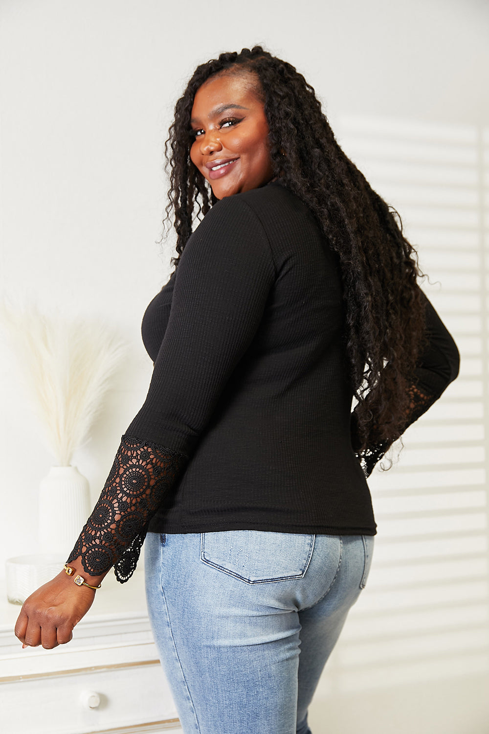 A woman with long curly hair wears the Double Take Crochet Lace Hem Sleeve Button Top and versatile solid color blue jeans, standing indoors with a neutral expression.