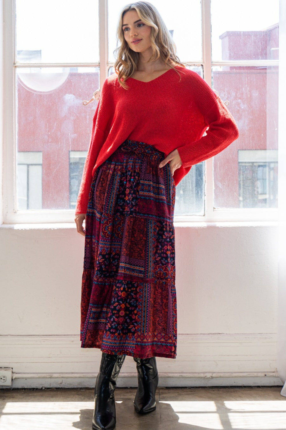 A person stands near a window exuding bohemian charm in a red sweater, an ODDI Full Size Printed Tiered Maxi Skirt, and black boots.