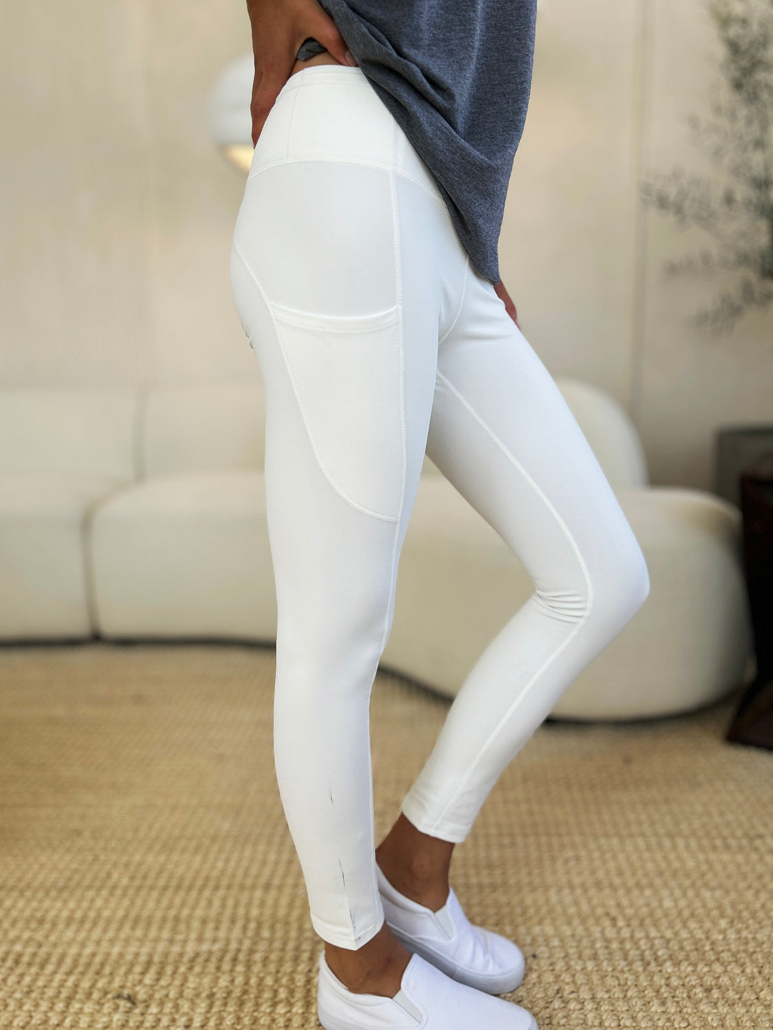 Person wearing Wide Waistband Sports Leggings made of stretchy nylon spandex and white slip-on shoes, standing on a beige carpet with a white couch in the background.