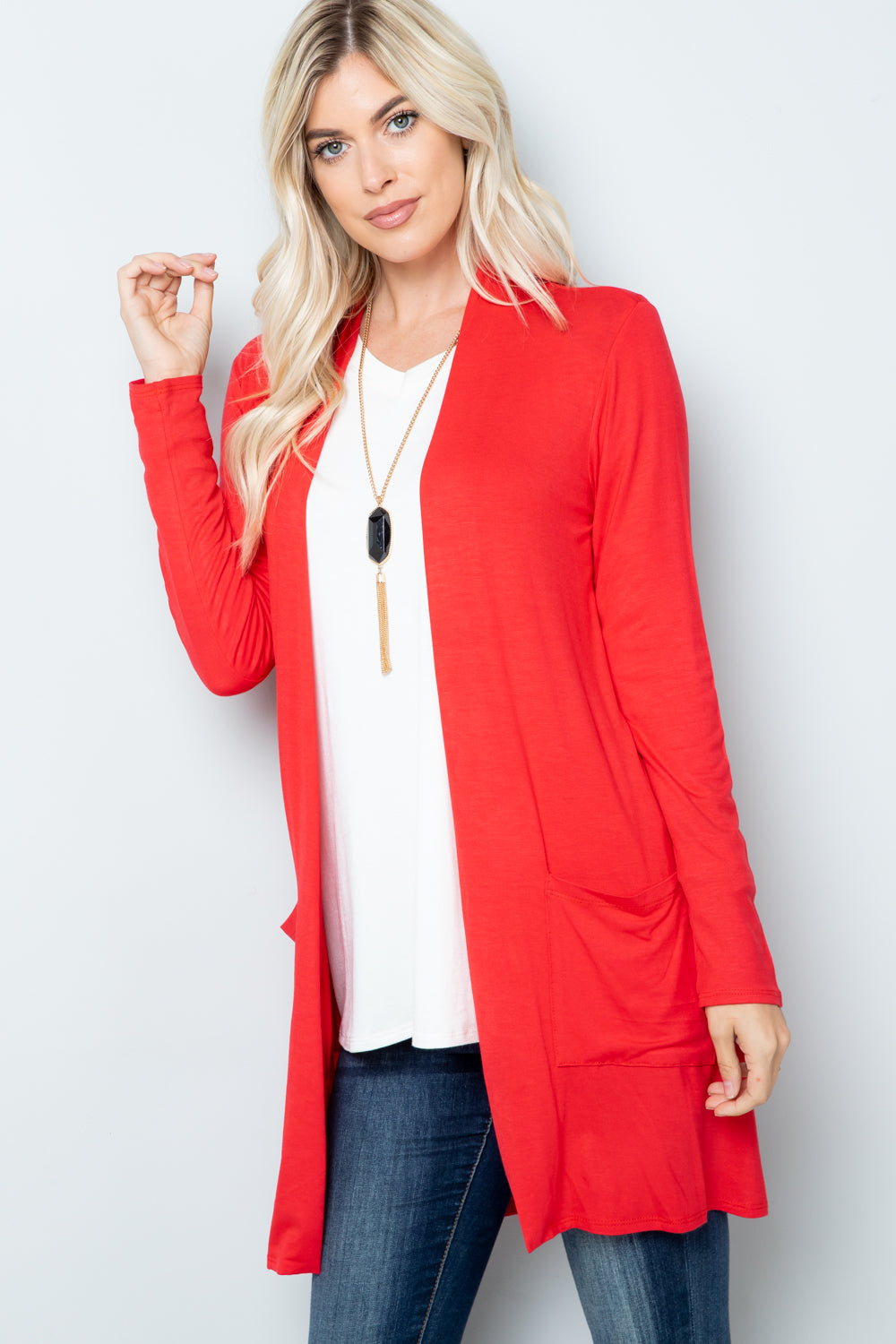 A woman in a highly stretchy Celeste Full Size Open Front Cardigan with Pockets, along with a white top and blue jeans, stands against a plain background, with one hand in her cardigan pocket, wearing a long necklace.