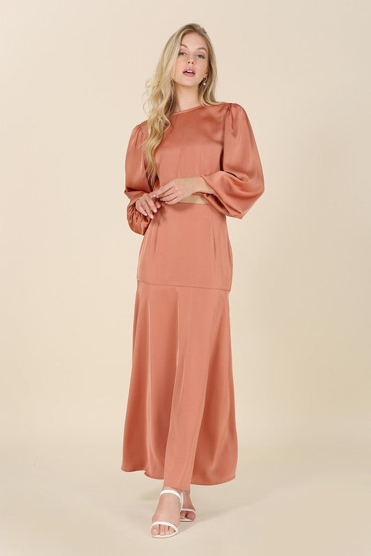 A woman stands against a plain background, elegantly adorned in the "Dressed up satin two-piece mermaid dress set" featuring a peach-colored, balloon-sleeve crop top and matching high-waisted maxi skirt, accessorized with white high-heeled sandals.