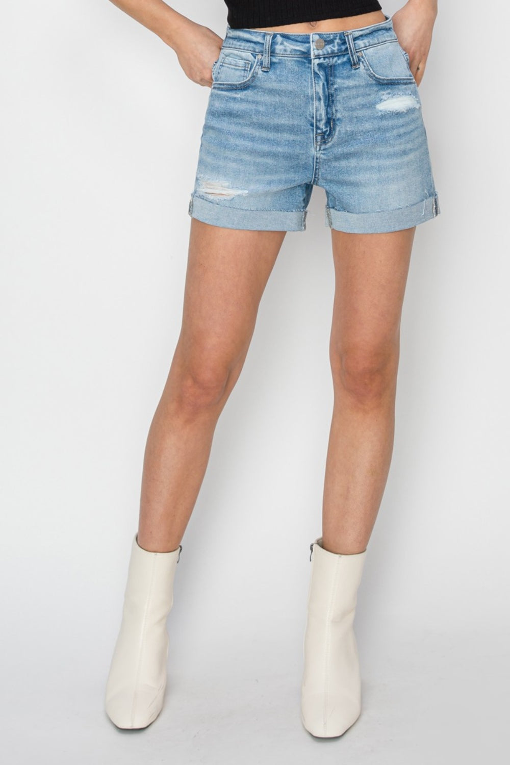 A person wearing RISEN distressed mid-rise waist denim shorts in light blue paired with white ankle boots and a black top, standing against a plain background—a staple for any summer wardrobe.