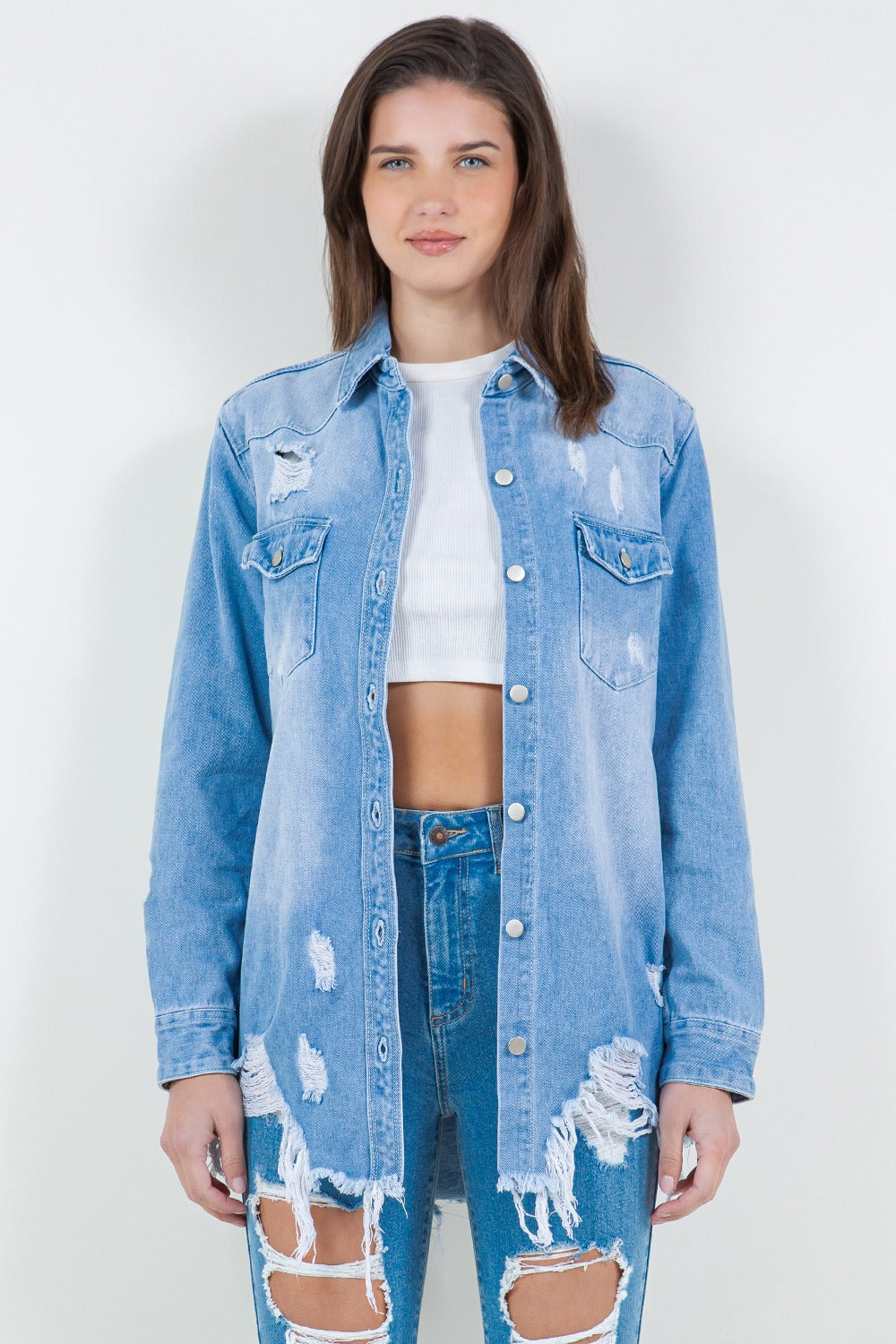 A woman stands against a plain background wearing the American Bazi Raw Hem Distressed Long Sleeve Denim Jacket over a white crop top and ripped jeans, adding an edgy flair to her wardrobe.