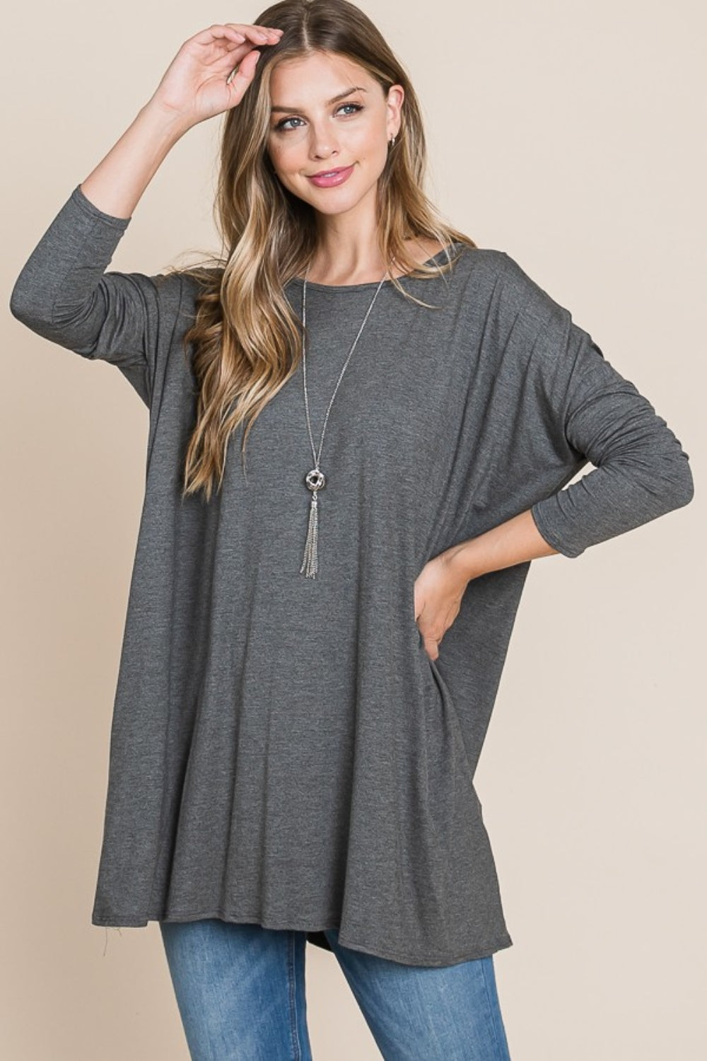 A woman with long hair is wearing a cozy, BOMBOM Round Neck Long Sleeve Oversized Top and blue jeans. She is smiling with one hand touching her head and the other in her pocket. Her comfortable chic wardrobe includes a long necklace, adding a stylish touch to her relaxed outfit.