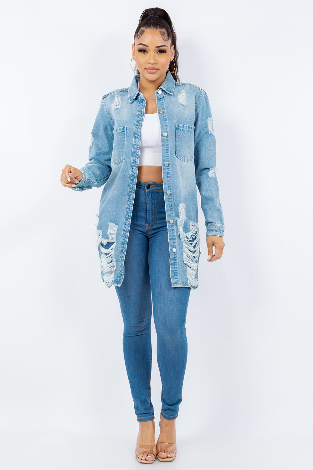 A woman with long hair is wearing an American Bazi Distressed Button Up Long Sleeve Denim Jacket over a white crop top and blue jeans. She is facing forward and standing against a plain white background.