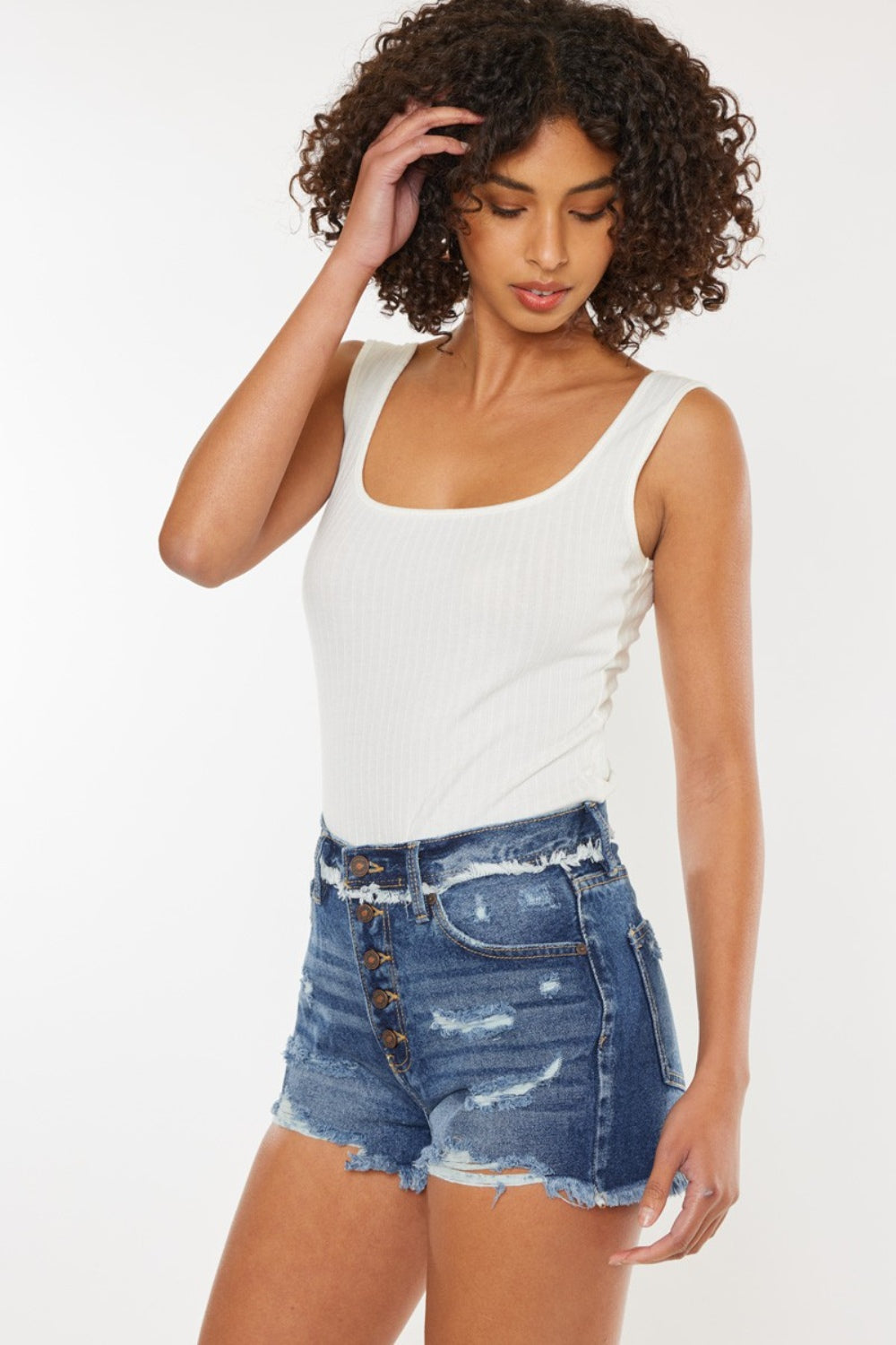 A woman with curly hair wearing a white sleeveless top and Kancan Distressed Button Fly Denim Shorts stands against a plain background, perfect for a stylish summer wardrobe.