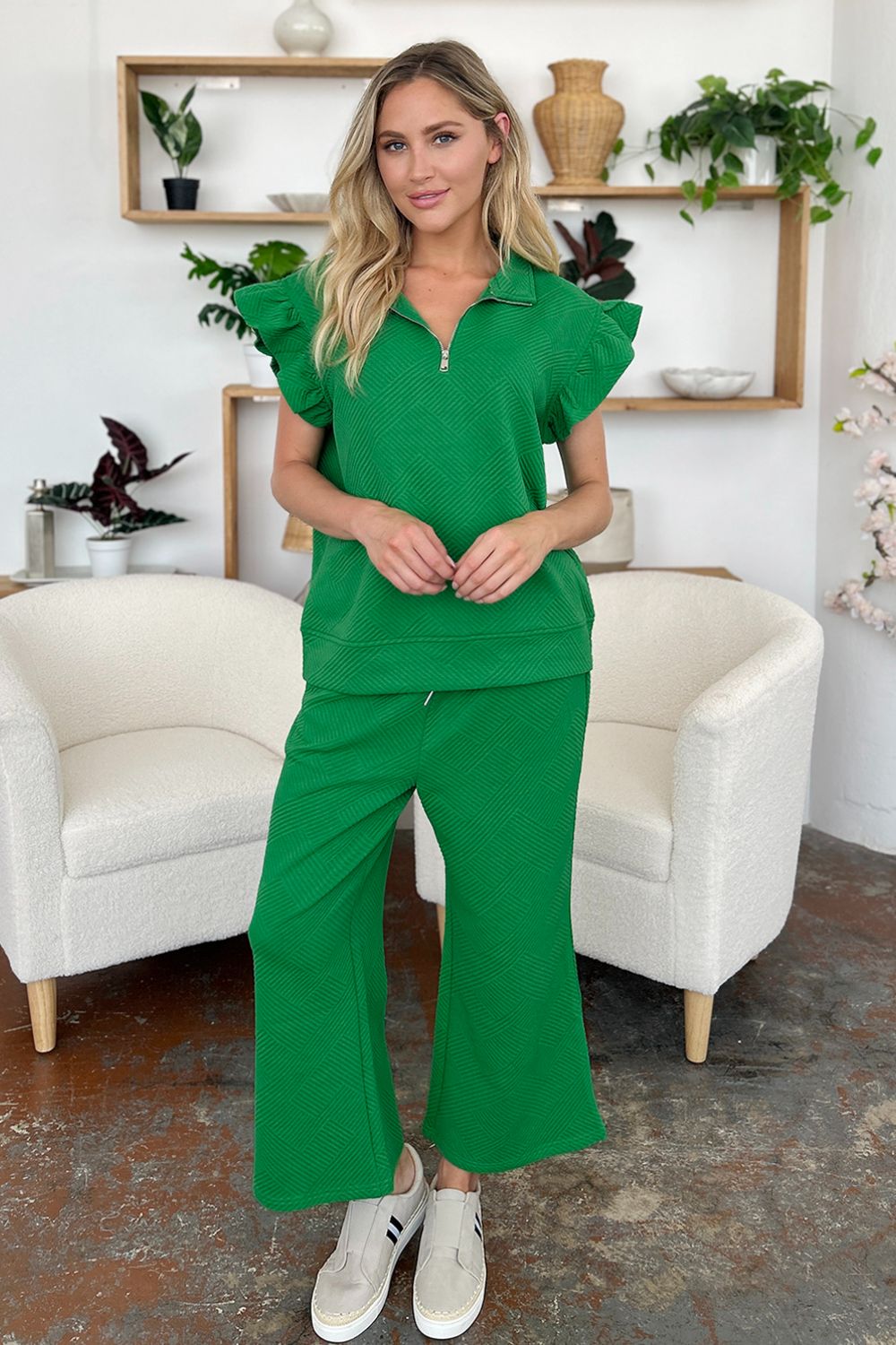 A woman stands wearing the Double Take Texture Ruffle Short Sleeve Top and Drawstring Wide Leg Pants Set in a slightly stretchy, light gray fabric with zipper detail. The two-piece outfit is complemented by black sandals, and she has one hand in her pocket. Machine wash cold for easy care.