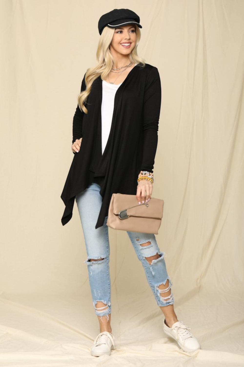 A woman wearing a black hat, the Celeste Full Size Open Front Knit Cardigan—an essential wardrobe staple—and a white top, holding a beige clutch, poses against a beige background.