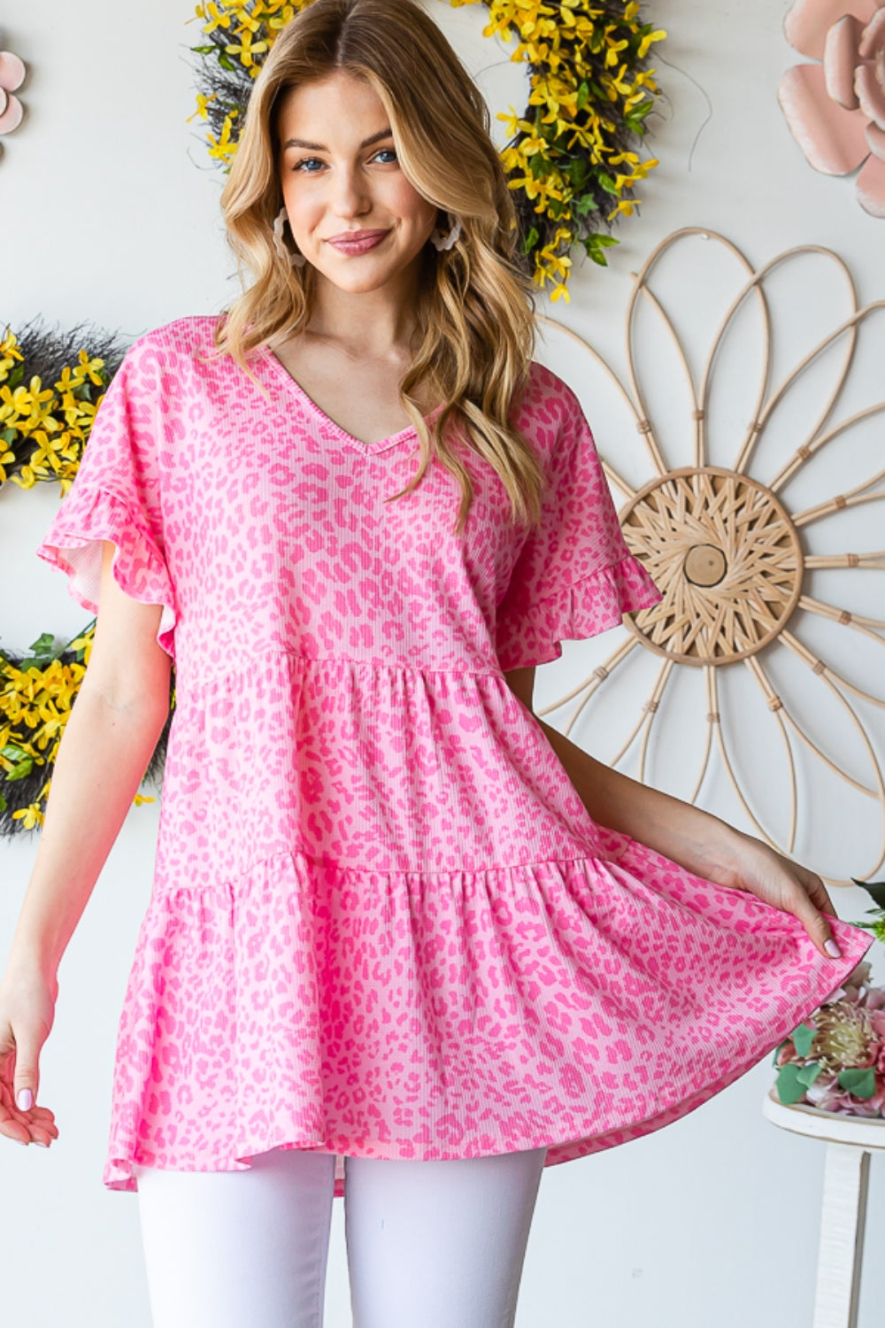 A person wearing a Heimish Full Size Leopard Ruffle Trim Short Sleeve Top stands in front of a floral wreath display.