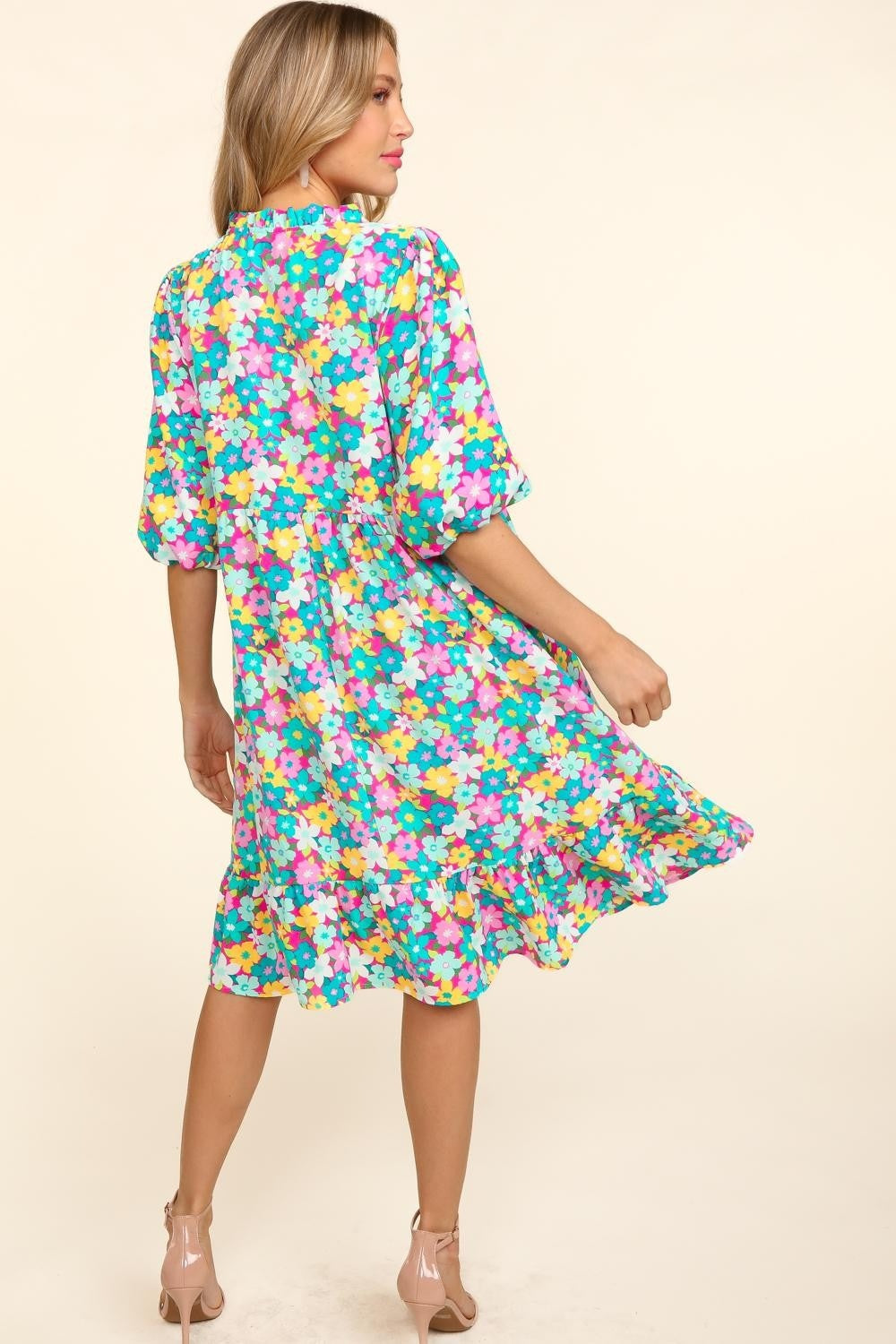 A woman in a Haptics Bubble Sleeve Floral Ruffled Dress stands against a plain background, with one hand holding her dress and the other by her side. She has long, wavy hair and is wearing open-toed shoes, perfect for any garden party.