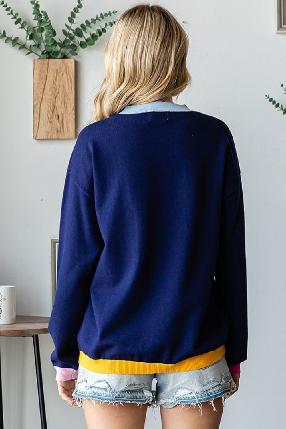 A person with long blonde hair wearing a First Love Contrast Ribbed Round Neck Long Sleeve Sweater and light denim shorts stands indoors near a small table and plant.