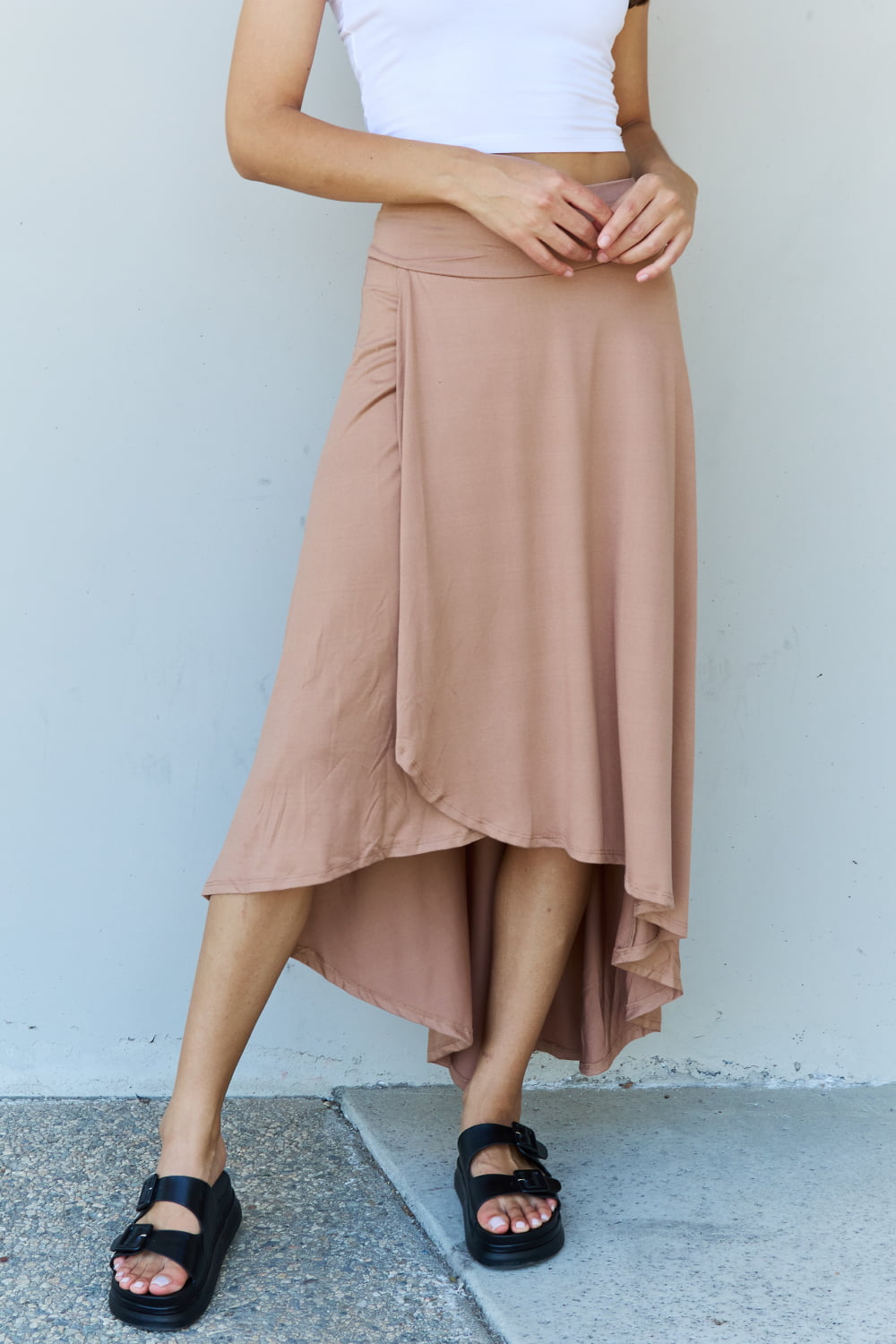 Person wearing a white crop top, the Ninexis First Choice High Waisted Flare Maxi Skirt in Camel with an asymmetrical hemline, and black sandals, standing against a light-colored wall.