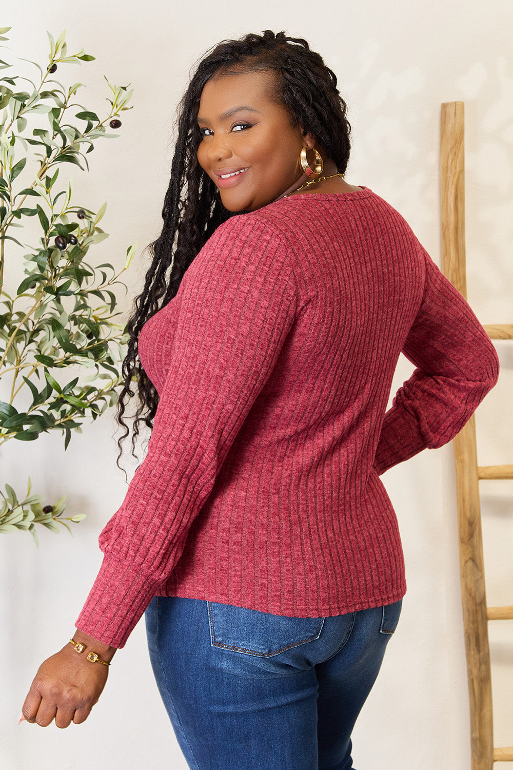 A person in a stylish maroon Ribbed Round Neck Lantern Sleeve Blouse and jeans stands by a wooden ladder, with olive branches in the background.