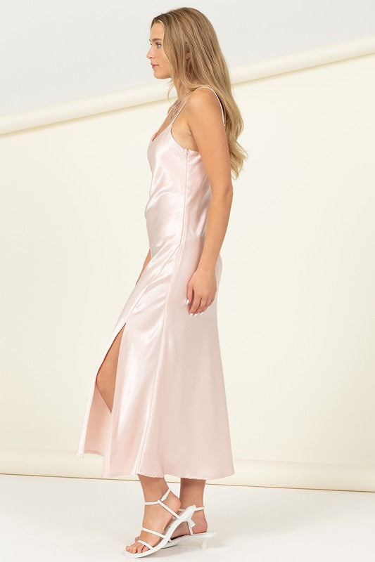 Dressed in the Love Tree Sleeveless Maxi Dress, a woman showcases effortless elegance as she holds the light pink satin skirt to reveal a thigh-high slit, set against a plain background.