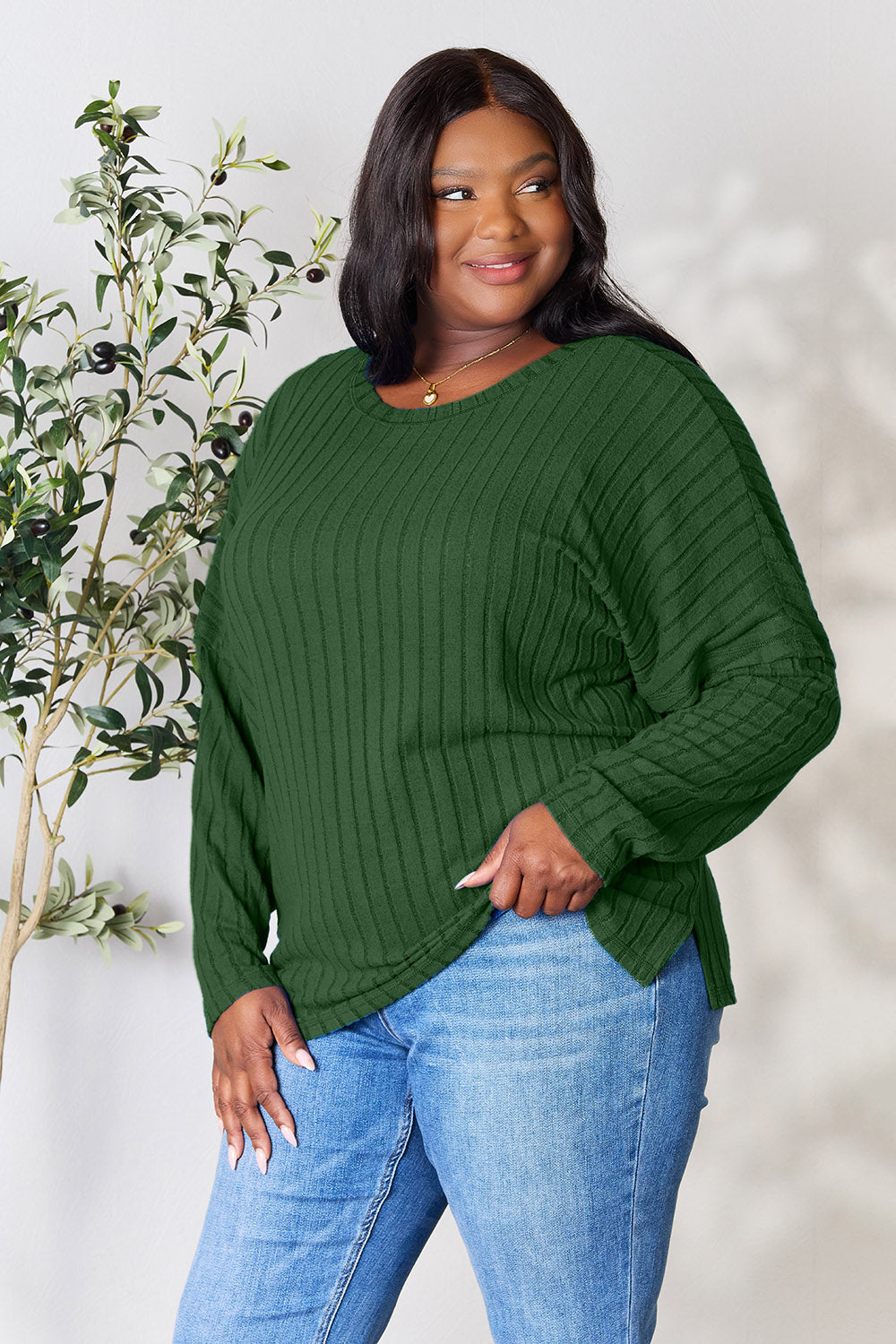 A person wearing the Basic Bae Full Size Ribbed Round Neck Slit Knit Top in blue and beige pants stands and smiles in front of a leafy plant.