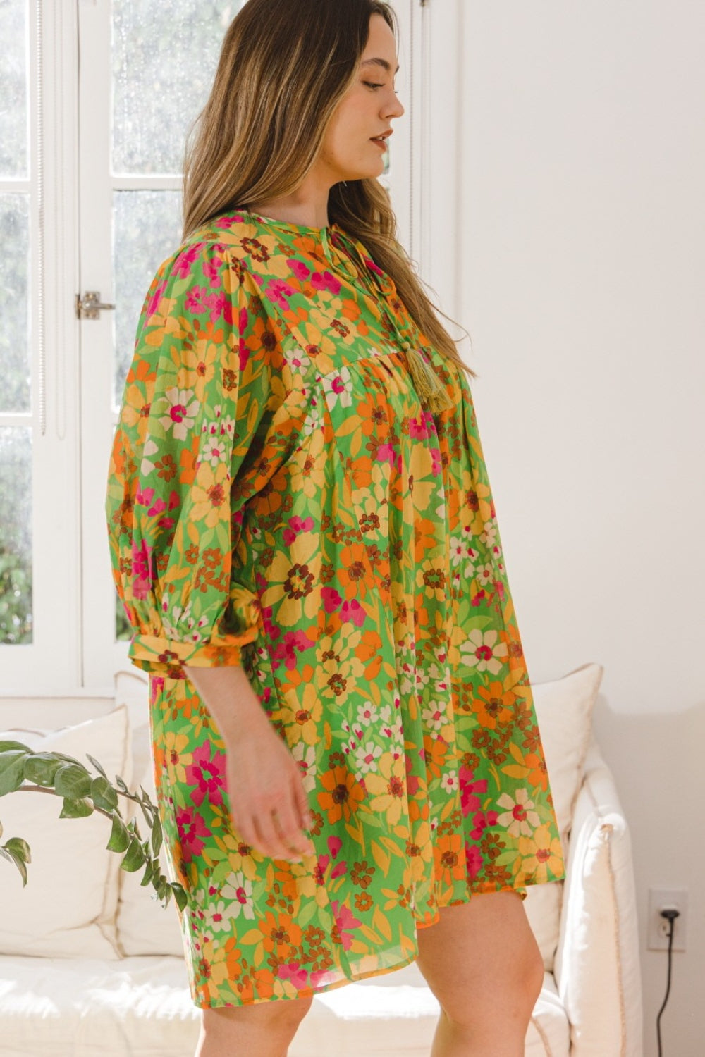 A person is seated on a beige couch, adorned in an ODDI Full Size Floral Tied Neck Mini Dress, beside a window that displays the lush greenery outside.